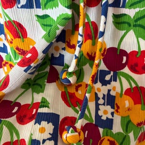 K&ouml;nnte beinhalten: Nahaufnahme eines wei&szlig;en Rippstoffs mit einem lebendigen Kirsch- und Blumenmuster in Rot, Gelb, Blau und Gr&uuml;n. Das Design umfasst dekorative Kordeln mit bunten geknoteten Enden.