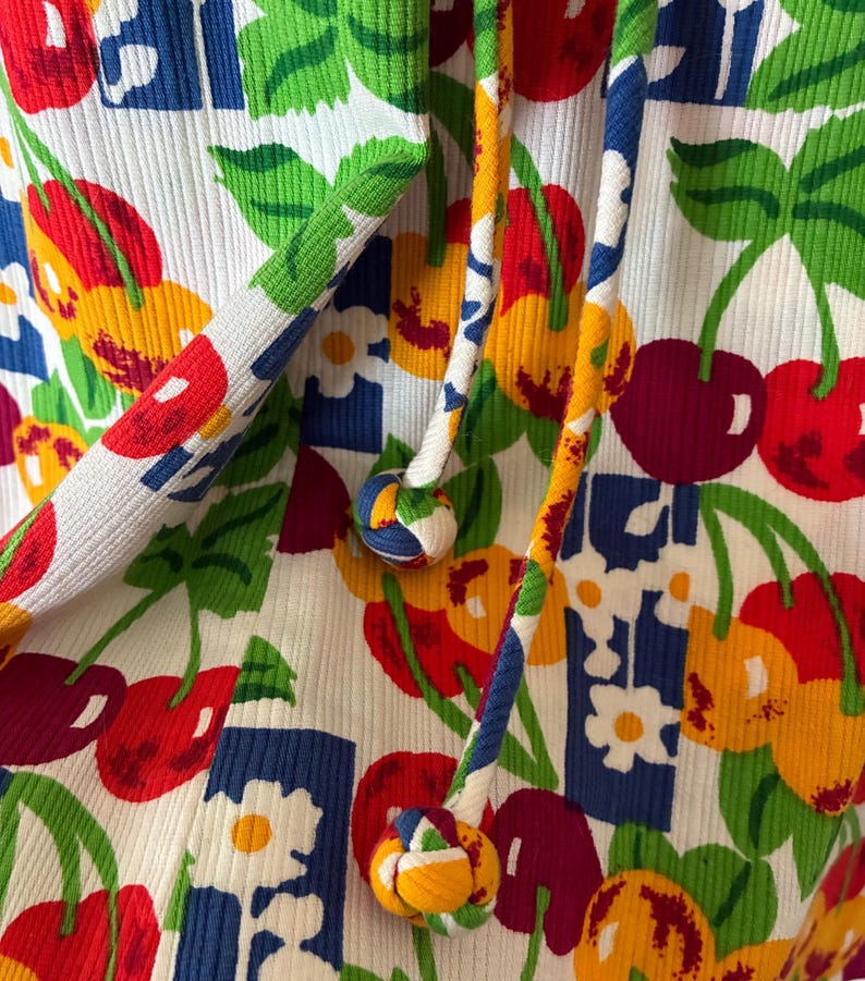 K&ouml;nnte beinhalten: Nahaufnahme eines wei&szlig;en Rippstoffs mit einem lebendigen Kirsch- und Blumenmuster in Rot, Gelb, Blau und Gr&uuml;n. Das Design umfasst dekorative Kordeln mit bunten geknoteten Enden.