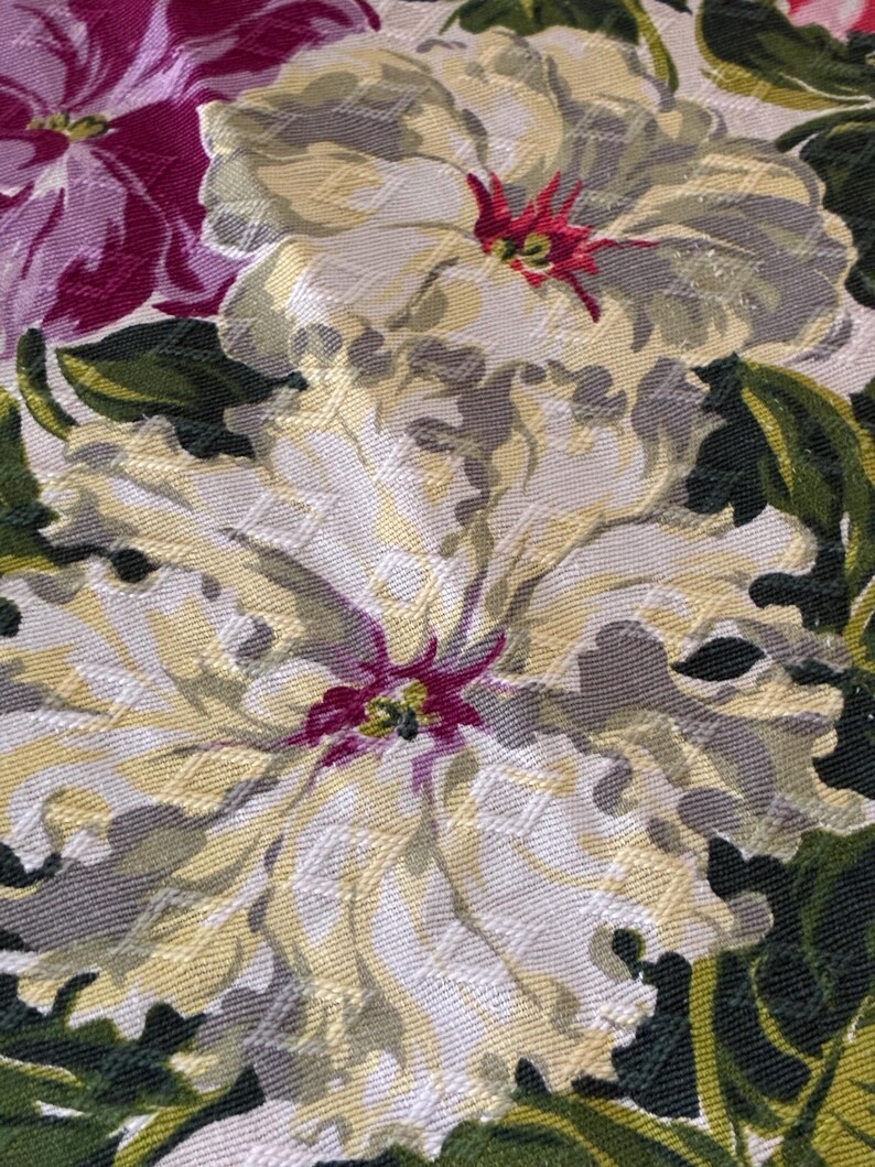 K&ouml;nnte beinhalten: Nahaufnahme eines floralen Stoffes mit gro&szlig;en, strukturierten Blumen in Creme-, Grau- und Gelbt&ouml;nen. Die Blumen haben rote und violette Zentren, umgeben von gr&uuml;nen Bl&auml;ttern. Der Stoff hat ein gewebtes Aussehen.