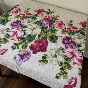 K&ouml;nnte beinhalten: Cremefarbene Tischdecke mit floralem Muster. Das Design zeigt gro&szlig;e Bl&uuml;ten in Rot-, Rosa-, Lila- und Wei&szlig;t&ouml;nen mit gr&uuml;nen Bl&auml;ttern. Der Stoff hat ein rautenf&ouml;rmiges Muster.