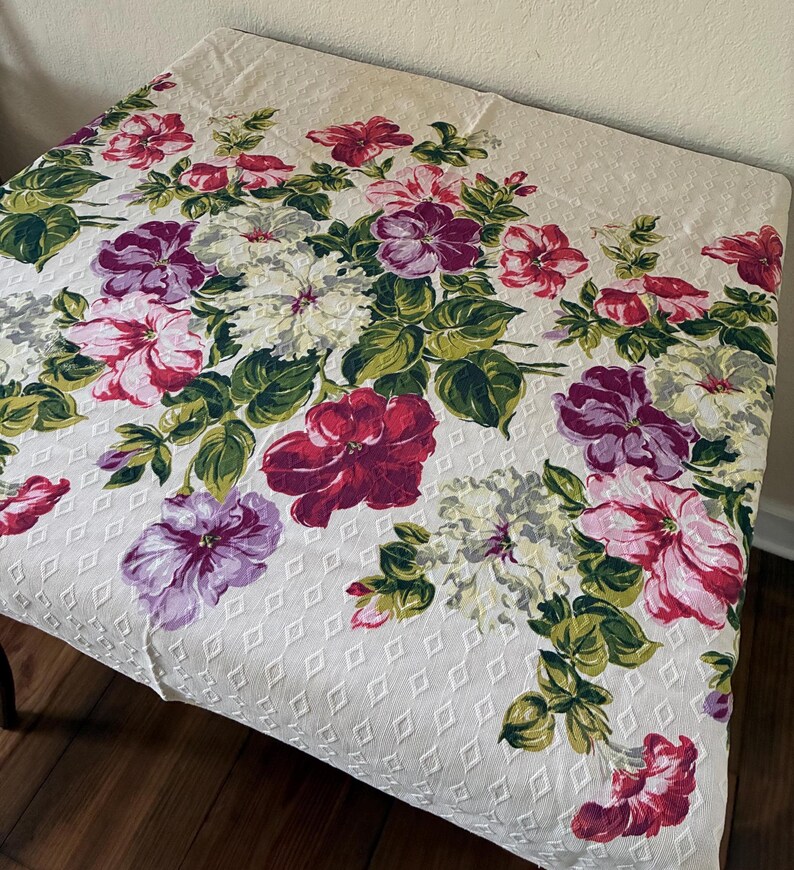 K&ouml;nnte beinhalten: Cremefarbene Tischdecke mit floralem Muster. Das Design zeigt gro&szlig;e Bl&uuml;ten in Rot-, Rosa-, Lila- und Wei&szlig;t&ouml;nen mit gr&uuml;nen Bl&auml;ttern. Der Stoff hat ein rautenf&ouml;rmiges Muster.