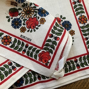 May include: White linen napkins with a floral design. The napkins feature a red border with green leaves and red berries. The corners are decorated with red roses, blue and yellow flowers, and the word "REINLEINEN".