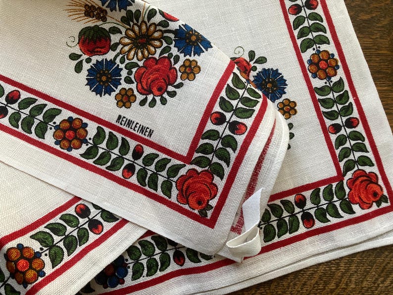 May include: White linen napkins with a floral design. The napkins feature a red border with green leaves and red berries. The corners are decorated with red roses, blue and yellow flowers, and the word "REINLEINEN".