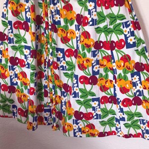 K&ouml;nnte beinhalten: Wei&szlig;er Stoff mit einem sich wiederholenden Muster aus roten und gelben Kirschen, gr&uuml;nen Bl&auml;ttern und kleinen wei&szlig;en Blumen. Die Kirschen sind auf einem Raster aus blauen Quadraten angeordnet. Der Stoff ist gerafft und erzeugt Falten und Schatten.