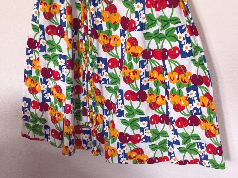 K&ouml;nnte beinhalten: Wei&szlig;er Stoff mit einem sich wiederholenden Muster aus roten und gelben Kirschen, gr&uuml;nen Bl&auml;ttern und kleinen wei&szlig;en Blumen. Die Kirschen sind auf einem Raster aus blauen Quadraten angeordnet. Der Stoff ist gerafft und erzeugt Falten und Schatten.