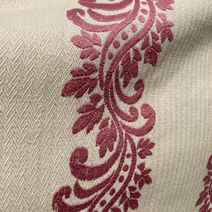 May include: A beige fabric featuring a woven herringbone texture and a prominent vertical band of deep magenta embroidery. The embroidery displays a continuous scrolling vine pattern with detailed leaves and small circular accents throughout.