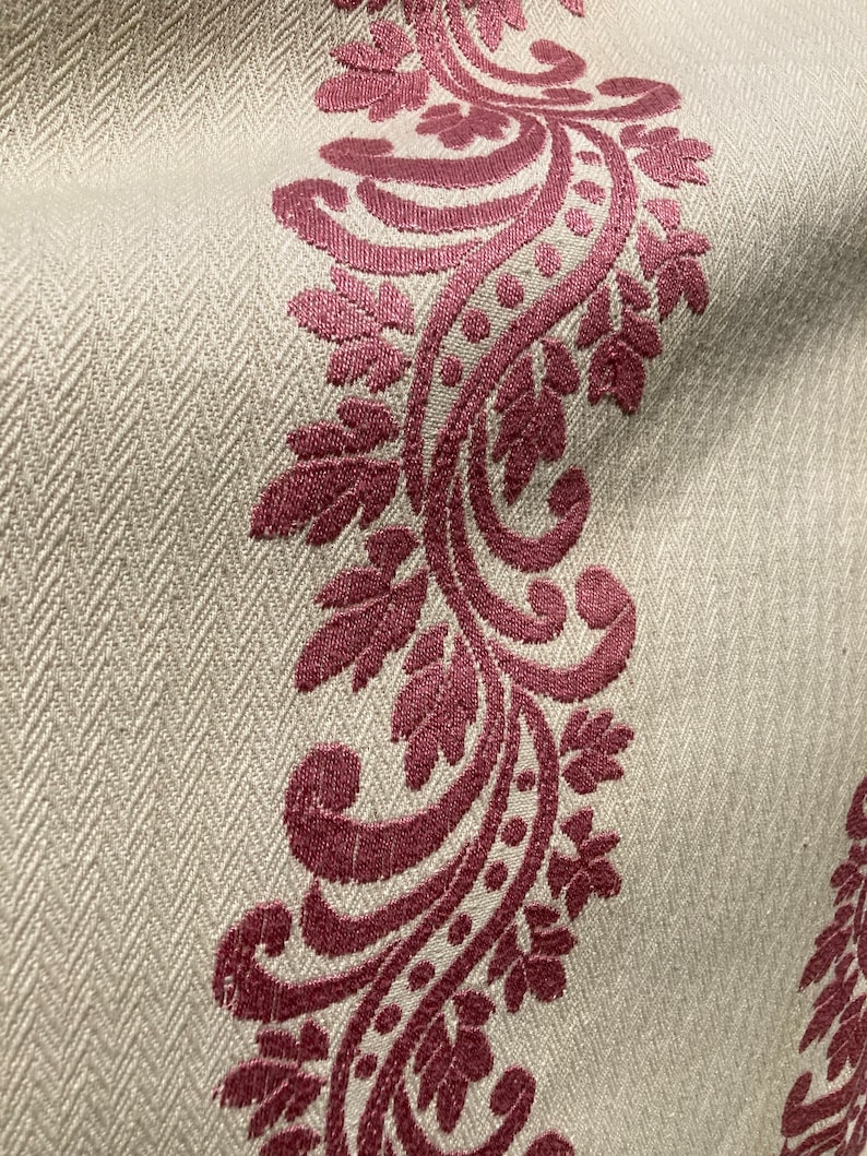 May include: A beige fabric featuring a woven herringbone texture and a prominent vertical band of deep magenta embroidery. The embroidery displays a continuous scrolling vine pattern with detailed leaves and small circular accents throughout.