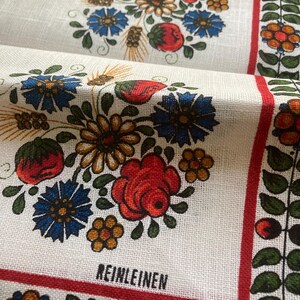 May include: White linen fabric with a floral design featuring red roses, blue flowers, and yellow accents. The fabric has a red border and the word "REINLEINEN" printed in black. The design is repeated across the fabric.