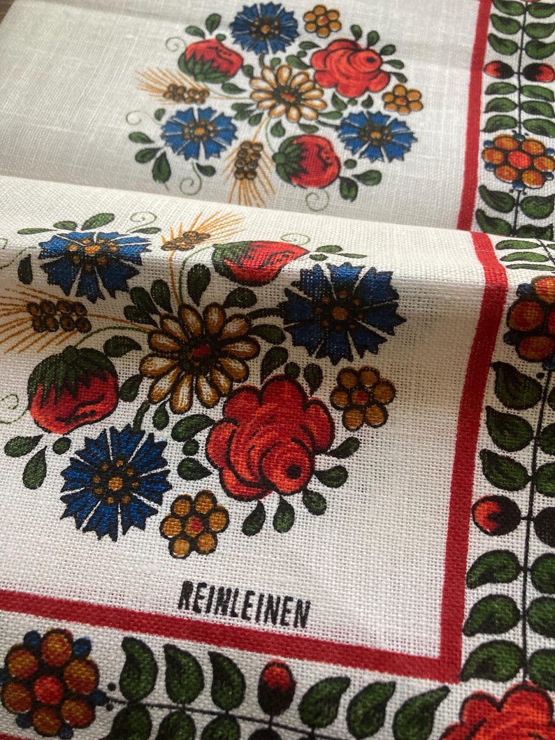 May include: White linen fabric with a floral design featuring red roses, blue flowers, and yellow accents. The fabric has a red border and the word "REINLEINEN" printed in black. The design is repeated across the fabric.
