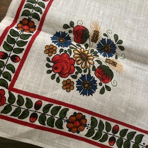 May include: White linen textile with a floral design. The border features a red stripe with green leaves and red berries. The center has a bouquet of red roses, blue flowers, and wheat stalks.