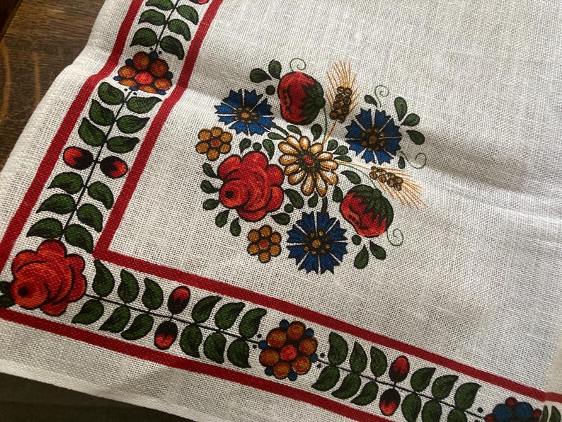 May include: White linen textile with a floral design. The border features a red stripe with green leaves and red berries. The center has a bouquet of red roses, blue flowers, and wheat stalks.
