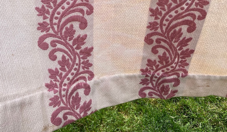 May include: A cream-colored fabric featuring a woven herringbone texture and vertical bands of raised, dark pink floral embroidery. The textile displays a repeating pattern of scrolling leaves and vines that run parallel across the surface.