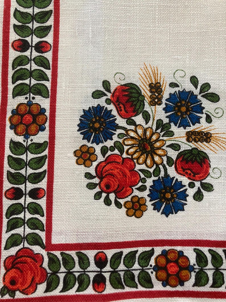 May include: A white fabric with a red and green border and a floral design in the corner. The floral design includes red, yellow, blue, and green flowers.