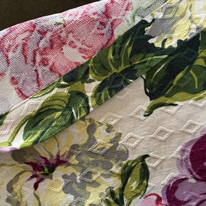 K&ouml;nnte beinhalten: Nahaufnahme eines floralen Stoffes mit cremefarbenem Hintergrund und Rautenmuster. Der Stoff zeigt gro&szlig;e Bl&uuml;ten in Rosa-, Lila-, Gelb- und Gr&uuml;nt&ouml;nen. Die Bl&auml;tter sind tiefgr&uuml;n.