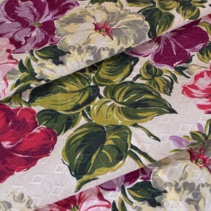 K&ouml;nnte beinhalten: Nahaufnahme eines floralen Stoffes mit gro&szlig;en, farbenfrohen Bl&uuml;ten. Die Blumen sind in Rot-, Rosa-, Lila- und Cremet&ouml;nen gehalten, mit gr&uuml;nen Bl&auml;ttern. Der Hintergrund ist cremefarben mit einem dezenten Rautenmuster. Der Stoff ist gefaltet und zeigt seine Textur.