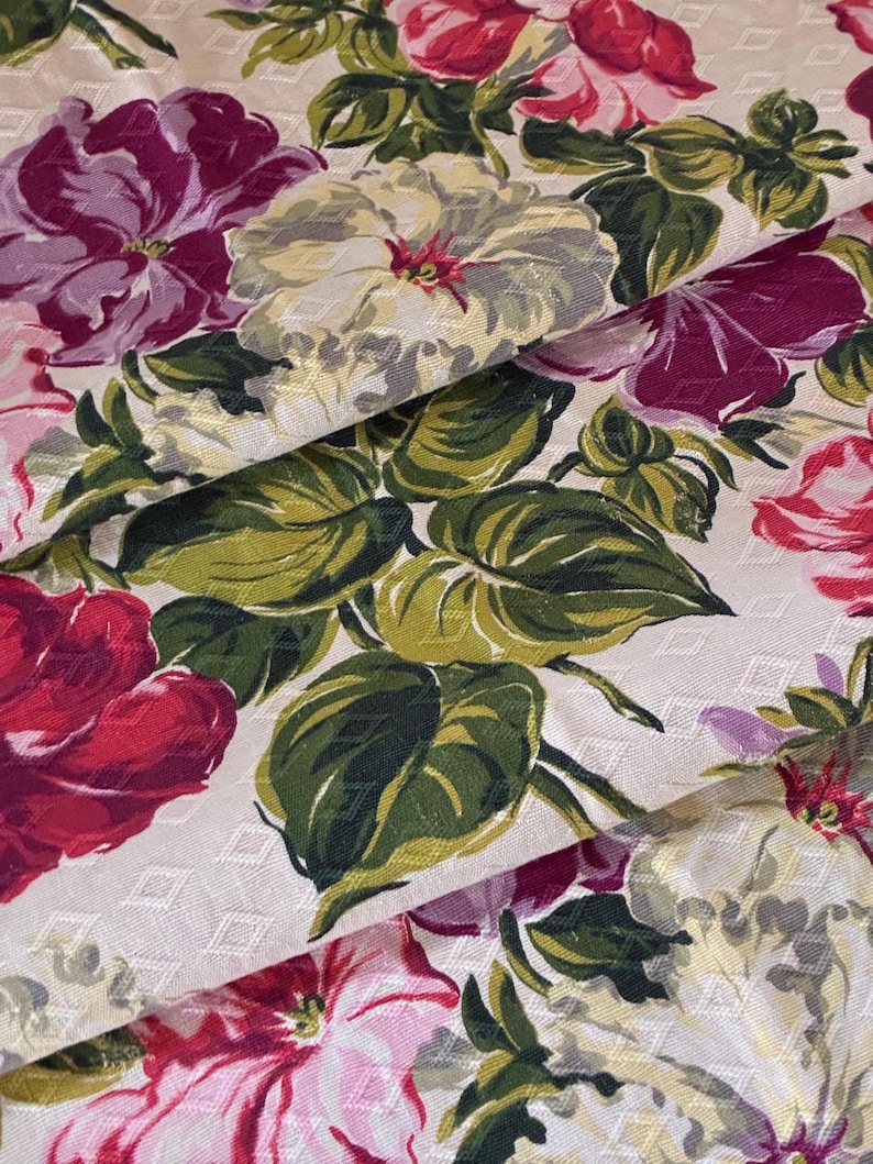 K&ouml;nnte beinhalten: Nahaufnahme eines floralen Stoffes mit gro&szlig;en, farbenfrohen Bl&uuml;ten. Die Blumen sind in Rot-, Rosa-, Lila- und Cremet&ouml;nen gehalten, mit gr&uuml;nen Bl&auml;ttern. Der Hintergrund ist cremefarben mit einem dezenten Rautenmuster. Der Stoff ist gefaltet und zeigt seine Textur.