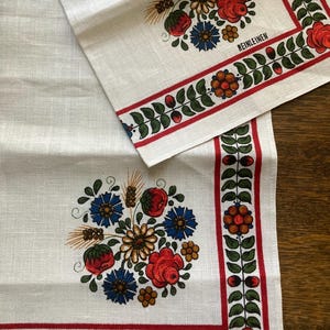 May include: White linen table runner and napkin set with a floral design. The design features red roses, blue flowers, and green leaves, with a red border. The word "REINLEINEN" is printed on the napkin.