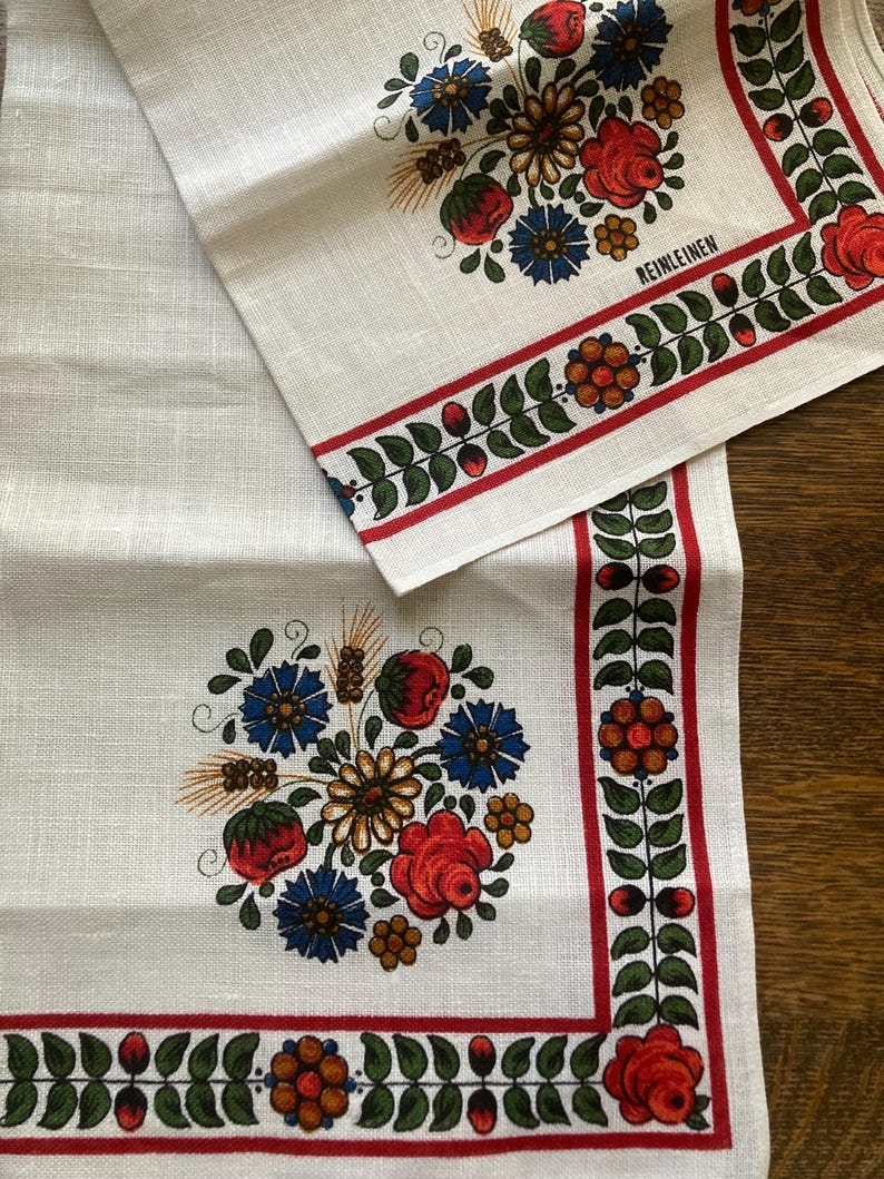 May include: White linen table runner and napkin set with a floral design. The design features red roses, blue flowers, and green leaves, with a red border. The word "REINLEINEN" is printed on the napkin.