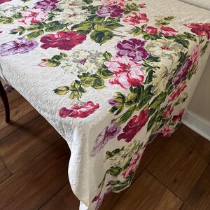 K&ouml;nnte beinhalten: Ein wei&szlig;es Tischtuch mit einem Blumenmuster, das gro&szlig;e Bl&uuml;ten in Rosa, Lila und Rot zeigt, akzentuiert durch gr&uuml;ne Bl&auml;tter. Das Tischtuch hat eine strukturierte, rautenf&ouml;rmige Oberfl&auml;che.