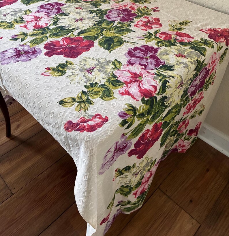 K&ouml;nnte beinhalten: Ein wei&szlig;es Tischtuch mit einem Blumenmuster, das gro&szlig;e Bl&uuml;ten in Rosa, Lila und Rot zeigt, akzentuiert durch gr&uuml;ne Bl&auml;tter. Das Tischtuch hat eine strukturierte, rautenf&ouml;rmige Oberfl&auml;che.