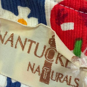 K&ouml;nnte beinhalten: Nahaufnahme eines cremefarbenen Stoffetiketts mit dem Schriftzug "Nantucket Naturals" in braunen Buchstaben. Das Etikett hat ein dekoratives Emblem. Der Hintergrund zeigt ein blau-wei&szlig;es Blumenmuster und ein rot-gr&uuml;nes Fruchtdesign.