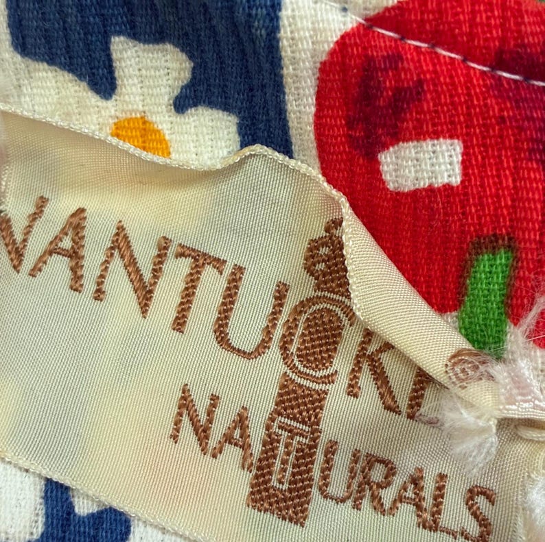 K&ouml;nnte beinhalten: Nahaufnahme eines cremefarbenen Stoffetiketts mit dem Schriftzug "Nantucket Naturals" in braunen Buchstaben. Das Etikett hat ein dekoratives Emblem. Der Hintergrund zeigt ein blau-wei&szlig;es Blumenmuster und ein rot-gr&uuml;nes Fruchtdesign.