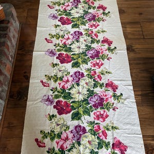K&ouml;nnte beinhalten: Cremefarbener Stoff mit einem Blumenmuster aus roten, rosa, lila und wei&szlig;en Bl&uuml;ten. Gr&uuml;ne Bl&auml;tter und Stiele sind mit den Bl&uuml;ten verwoben. Der rechteckige Stoff liegt auf einer Holzoberfl&auml;che.