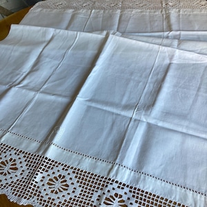 May include: White linen pillowcases with intricate, openwork lace detailing along the edges. The pillowcases are folded, showcasing the delicate floral and geometric patterns of the lace. The fabric appears crisp and clean.