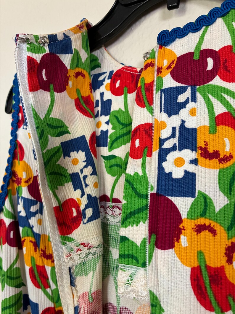 K&ouml;nnte beinhalten: Ein Kleid im Vintage-Stil mit einem lebendigen Kirschdruck in Rot, Gelb und Lila auf cremefarbenem Hintergrund. Das Kleid hat blaue Verzierungen, einen Rei&szlig;verschluss und wei&szlig;e Spitzendetails. Der Stoff hat eine gerippte Textur.