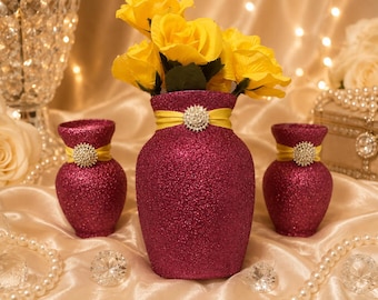 May include: A set of three magenta glitter-covered vases, each featuring a yellow ribbon band around the neck adorned with a circular silver-toned rhinestone embellishment. The central vase contains a bouquet of bright yellow artificial roses.