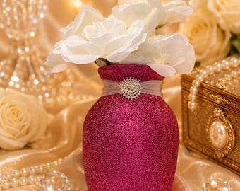 May include: A bright magenta glitter vase with a rounded body and a narrow neck. A sheer white ribbon is wrapped around the neck, secured by a circular silver-toned brooch featuring clear rhinestones. The vase contains several white silk roses.