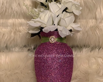 May include: A decorative vase with a glittery magenta finish, adorned with a green ribbon and a sparkling brooch. The vase holds a bouquet of white roses. The text "@exotiques creations" is visible on the vase.