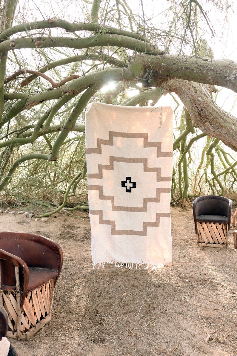 Op de afbeelding: Een wit geweven tapijt met een bruin geometrisch patroon hangt aan een tak. Het tapijt heeft een franje rand en een zwart kruis in het midden. Twee bruine rieten stoelen staan op de achtergrond.