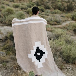May include: A beige woven blanket with white stripes and a geometric black and white design. The blanket is draped over a person in a natural outdoor setting, with desert plants in the foreground.