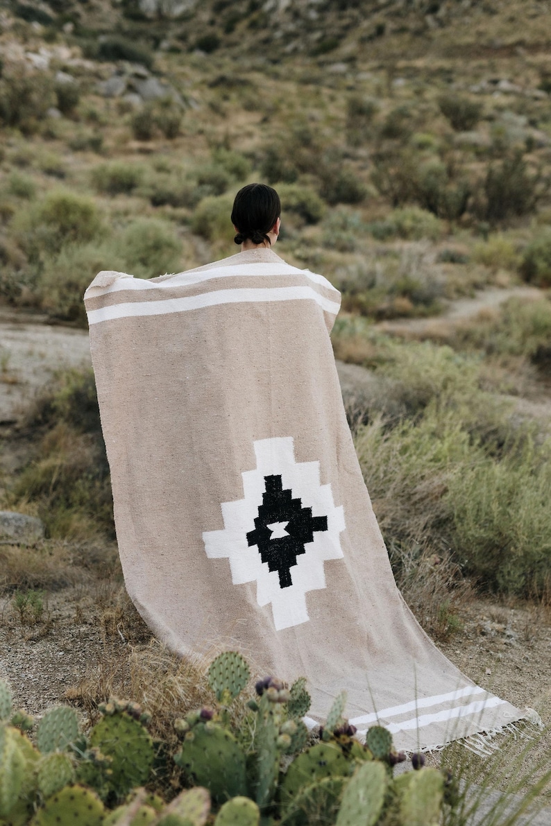 May include: A beige woven blanket with white stripes and a geometric black and white design. The blanket is draped over a person in a natural outdoor setting, with desert plants in the foreground.