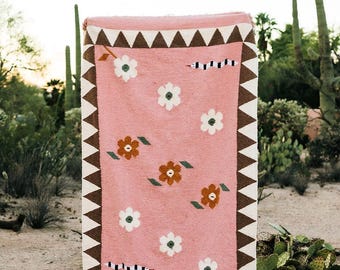May include: A pink woven rug with a floral pattern and a geometric border. The rug features white and brown triangles along the edges, with white flowers and brown flowers with green leaves scattered across the surface. A black and white striped design is also present.