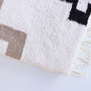 Op de afbeelding: Een wit geweven tapijt met bruine en zwarte geometrische patronen. Het tapijt heeft franjes aan de randen.