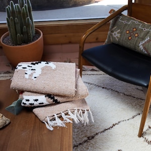 May include: A stack of beige woven blankets with a black and white horse design. The blankets have fringe along the bottom edge and are folded on a wooden table. A cactus in a terracotta pot is in the background.