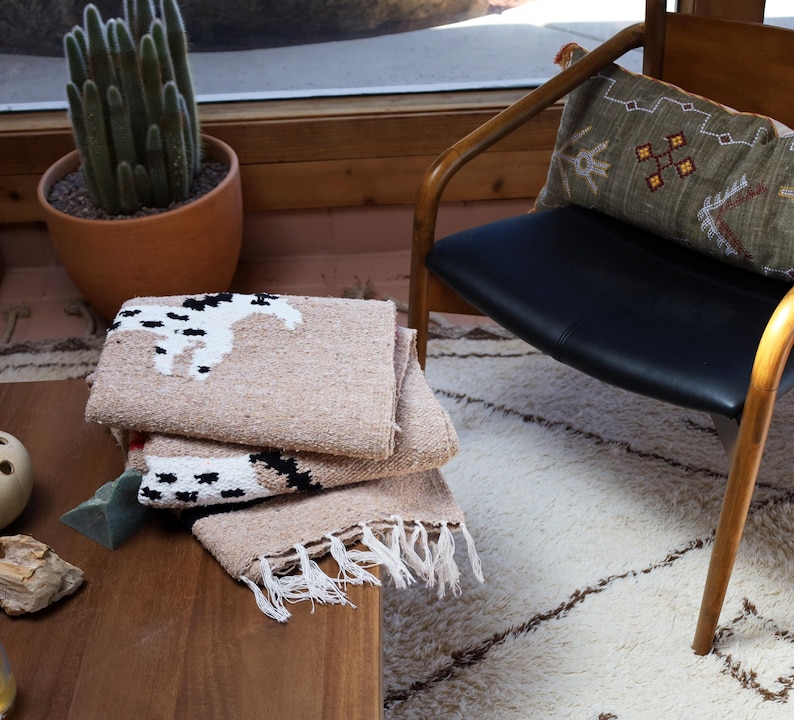May include: A stack of beige woven blankets with a black and white horse design. The blankets have fringe along the bottom edge and are folded on a wooden table. A cactus in a terracotta pot is in the background.