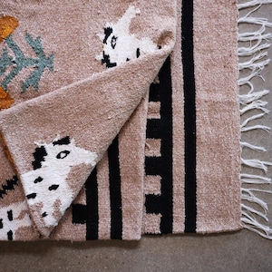 May include: A tan woven textile with black and white animal-like figures and black vertical stripes. The textile has a fringed edge and folded corner, revealing the pattern. The design includes orange and teal floral-like shapes.