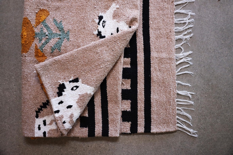 May include: A tan woven textile with black and white animal-like figures and black vertical stripes. The textile has a fringed edge and folded corner, revealing the pattern. The design includes orange and teal floral-like shapes.
