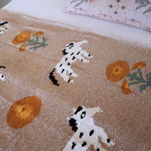 May include: A tan-colored textile featuring a repeating pattern of white and black spotted animal figures and orange floral designs. The textile is likely a rug or blanket, with a textured surface. A decorative pillow is visible in the background.