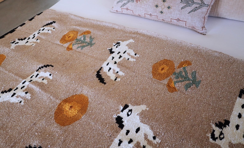 May include: A tan-colored textile featuring a repeating pattern of white and black spotted animal figures and orange floral designs. The textile is likely a rug or blanket, with a textured surface. A decorative pillow is visible in the background.