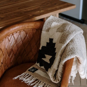 May include: A beige and black patterned blanket with fringe is draped over a brown leather chair. The blanket features a geometric design and a small label. The chair has a woven back and a rounded seat.
