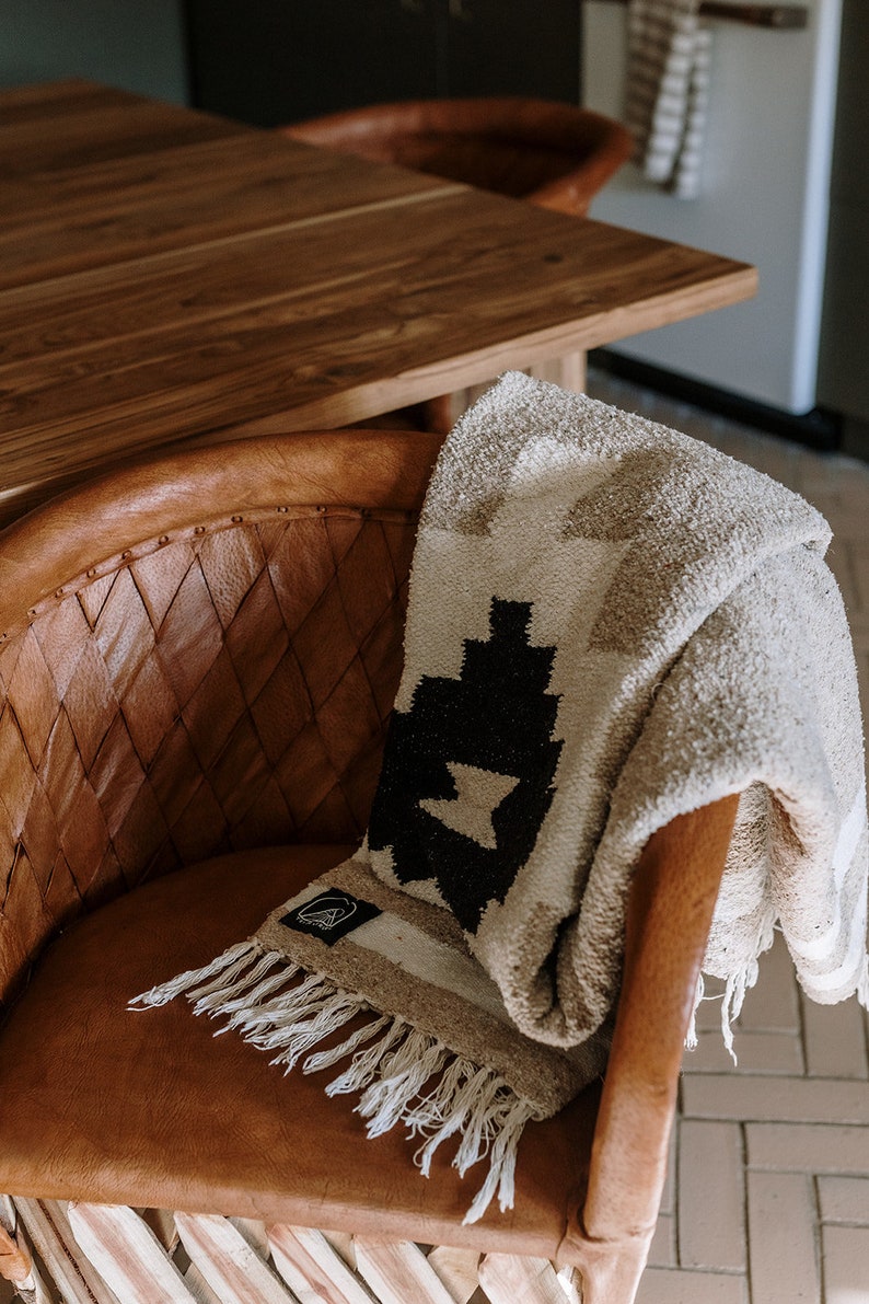 May include: A beige and black patterned blanket with fringe is draped over a brown leather chair. The blanket features a geometric design and a small label. The chair has a woven back and a rounded seat.