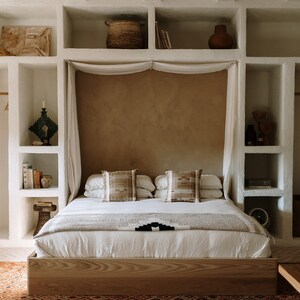 May include: A bedroom interior featuring a wooden bed frame, white bedding, and decorative pillows. The bed is centered against a textured wall, with built-in shelving on either side. A woven basket, books, and a robe are visible.