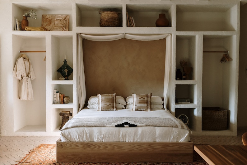 May include: A bedroom interior featuring a wooden bed frame, white bedding, and decorative pillows. The bed is centered against a textured wall, with built-in shelving on either side. A woven basket, books, and a robe are visible.
