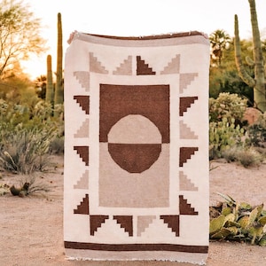 May include: A rectangular woven blanket with a geometric design in shades of brown, beige, and white. The blanket features a central circle and square pattern, with triangular accents. The blanket is displayed outdoors.