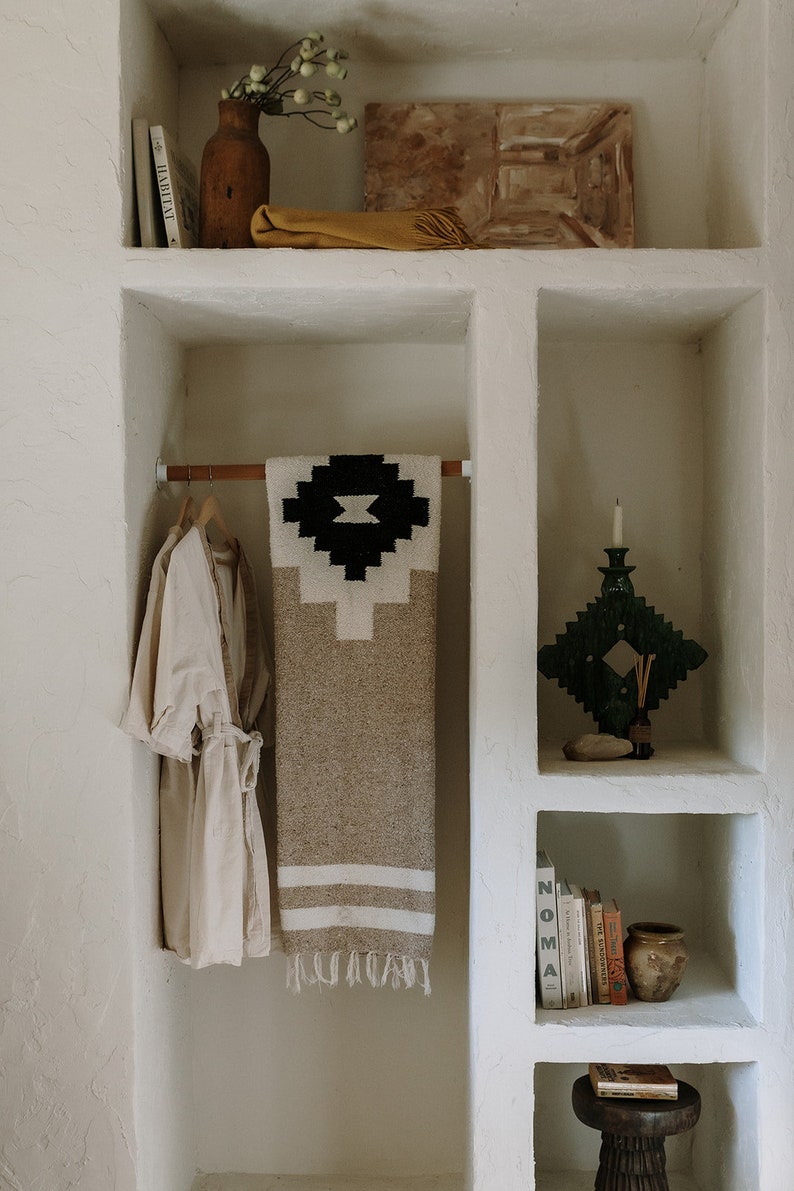 May include: A neutral-toned, built-in shelving unit with decorative items. A beige robe hangs on a wooden rod, next to a patterned blanket. Shelves hold books, a candle holder, a vase, and other decor.