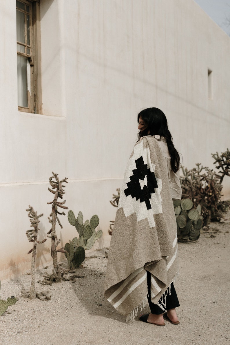 May include: A beige and white woven blanket with a geometric black design. The blanket is draped over a person's shoulders. The blanket has a fringed edge and is made of a soft material. The person is standing outside near cacti.